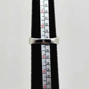 May include: A silver ring is placed on a black ring sizer. The ring is positioned at size 12. The ring sizer has measurements from 19 to 32. The ring is a simple band design.