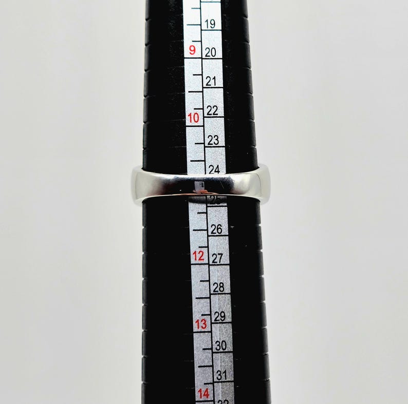 May include: A silver ring is placed on a black ring sizer. The ring is positioned at size 12. The ring sizer has measurements from 19 to 32. The ring is a simple band design.