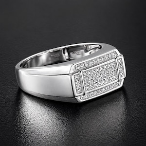 May include: A silver ring with a rectangular face, encrusted with small, sparkling stones. The ring has a polished finish and a wide band. The design features a central rectangle of stones, framed by smaller stone accents, creating a luxurious look.