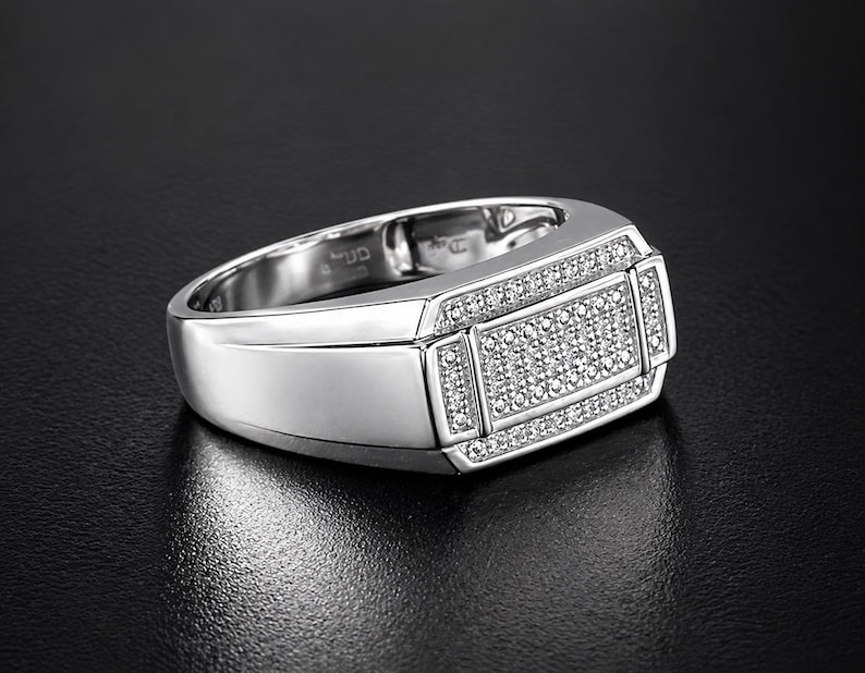 May include: A silver ring with a rectangular face, encrusted with small, sparkling stones. The ring has a polished finish and a wide band. The design features a central rectangle of stones, framed by smaller stone accents, creating a luxurious look.