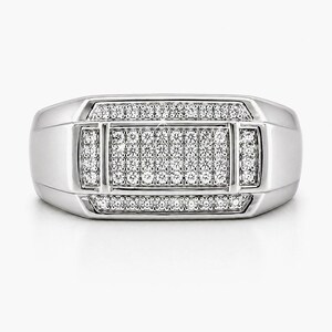May include: A silver-colored ring with a rectangular face, encrusted with numerous small, clear gemstones. The ring has a polished finish and a classic, geometric design. The gemstones are arranged in a symmetrical pattern, creating a sparkling effect.