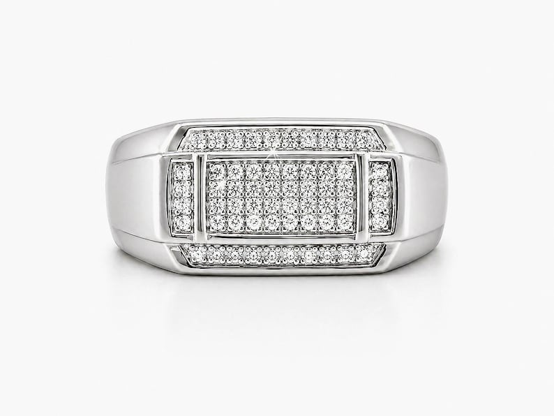 May include: A silver-colored ring with a rectangular face, encrusted with numerous small, clear gemstones. The ring has a polished finish and a classic, geometric design. The gemstones are arranged in a symmetrical pattern, creating a sparkling effect.