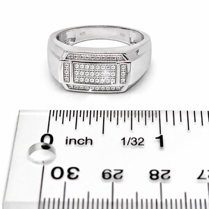 May include: A silver ring with a rectangular center stone, encrusted with small, clear stones. The ring has a wide band and is marked with "925". The ruler shows the ring is approximately 1 inch in diameter.