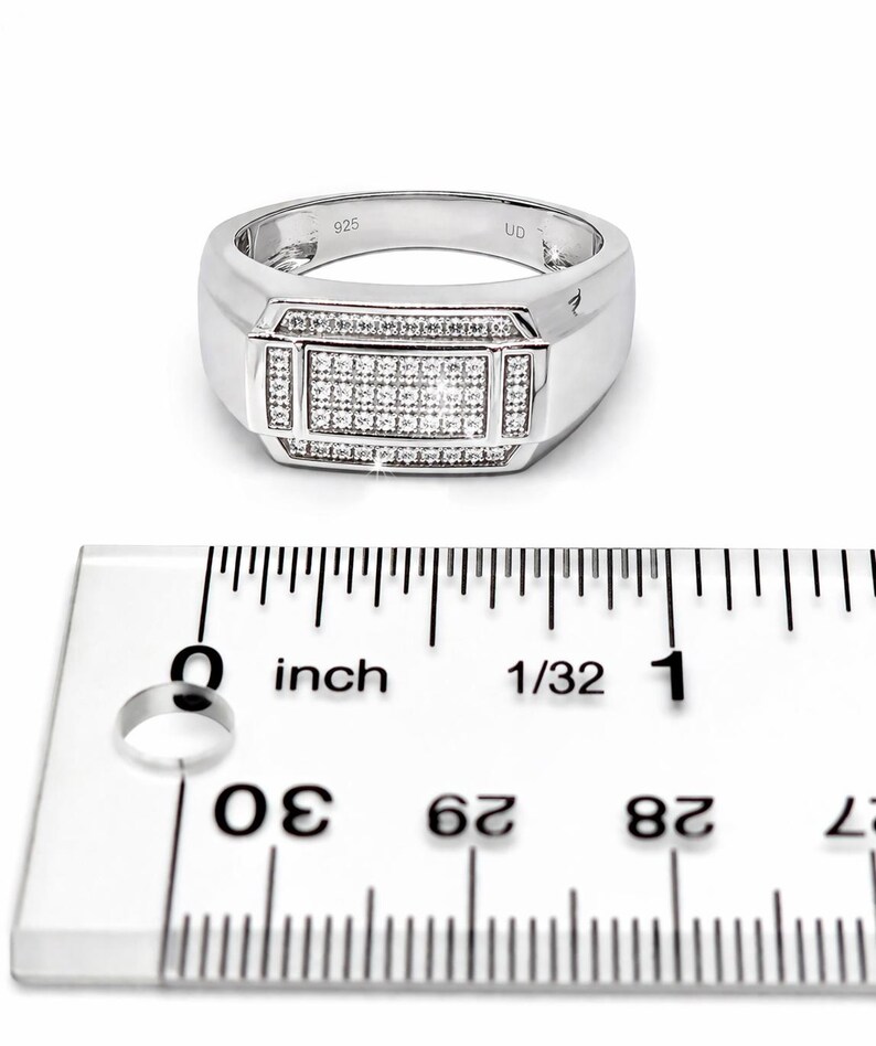 May include: A silver ring with a rectangular center stone, encrusted with small, clear stones. The ring has a wide band and is marked with "925". The ruler shows the ring is approximately 1 inch in diameter.