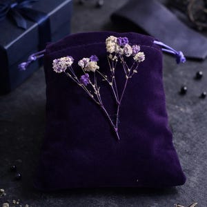 May include: A deep purple velvet drawstring bag is adorned with delicate dried flowers in white and purple. A navy blue gift box with a matching ribbon and a pair of silver cufflinks are also visible, suggesting a gift set.