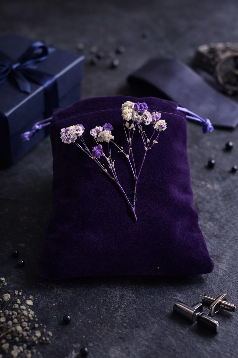 May include: A deep purple velvet drawstring bag is adorned with delicate dried flowers in white and purple. A navy blue gift box with a matching ribbon and a pair of silver cufflinks are also visible, suggesting a gift set.