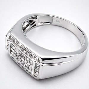 May include: A silver ring with a rectangular face, encrusted with small, sparkling stones. The band is smooth and polished, with the number "925" engraved on the inside. The ring has a modern, geometric design.