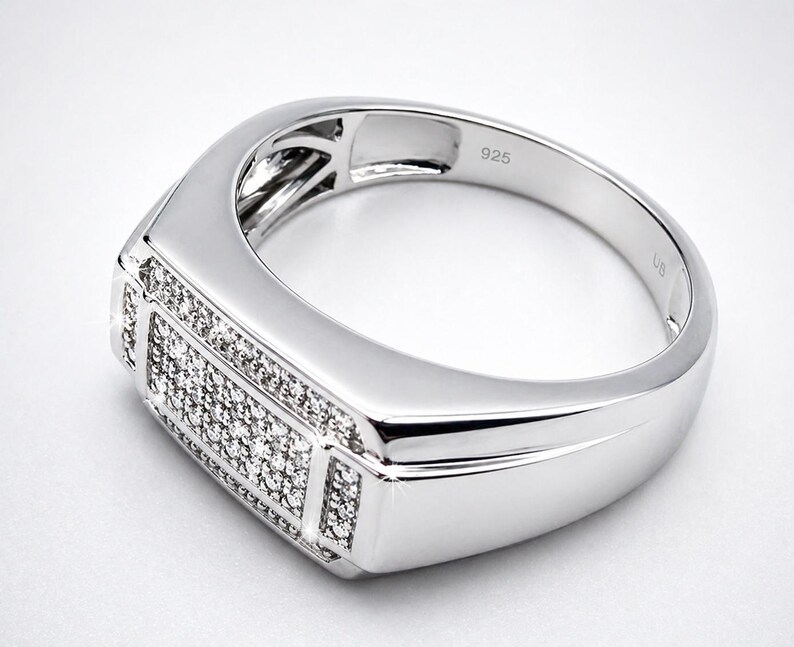 May include: A silver ring with a rectangular face, encrusted with small, sparkling stones. The band is smooth and polished, with the number "925" engraved on the inside. The ring has a modern, geometric design.