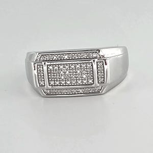 May include: A silver-toned ring with a rectangular center, encrusted with small, clear stones. The ring has a geometric design with additional stones bordering the central rectangle. The band is smooth and polished.
