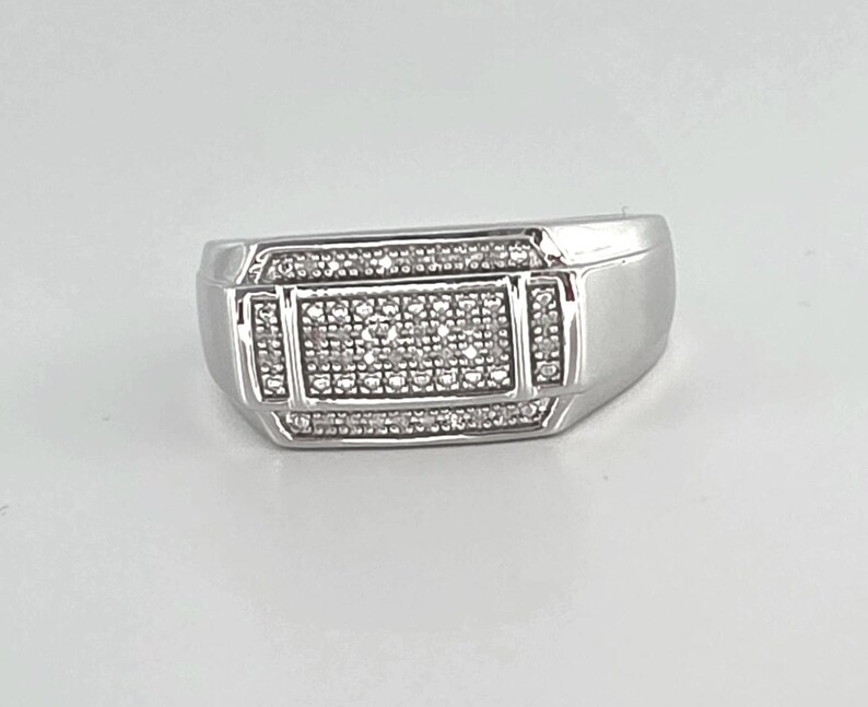 May include: A silver-toned ring with a rectangular center, encrusted with small, clear stones. The ring has a geometric design with additional stones bordering the central rectangle. The band is smooth and polished.