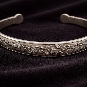 以下が含まれることがあります： オープンデザインのシルバーカフブレスレット。ブレスレットは、詳細な花と葉が特徴のフローラルパターンで飾られています。ジュエリーは、暗く質感のある表面に表示されています。