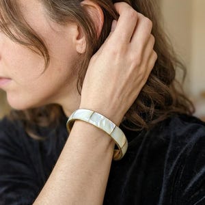 Pode incluir: Uma pulseira rígida articulada com segmentos retangulares de madrepérola branca iridescente incrustados numa estrutura de metal dourado polido. A pulseira tem um perfil curvo e liso, com um mecanismo de fecho seguro visível na lateral.