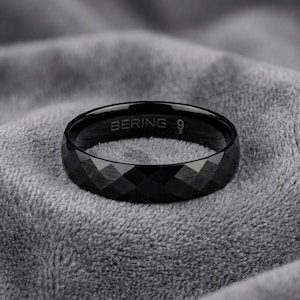Peut inclure: Une bague noire avec une texture de surface géométrique à facettes rappelant le diamant. L'intérieur de l'anneau est gravé avec le texte BERING et le chiffre 9. La bague présente une finition sombre et polie qui reflète la lumière sur son extérieur.