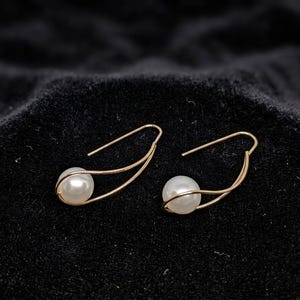 Puede incluir: Un par de pendientes de hilo dorado, cada uno con una perla esférica blanca engastada en un diseño de metal curvado de bucle abierto. Los pendientes cuentan con un cierre de gancho sencillo y un acabado pulido que ofrece un aspecto equilibrado.