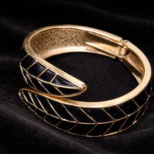 Puede incluir: Un brazalete dorado con un diseño geométrico negro. El brazalete con bisagras presenta un patrón repetido de formas de diamante negro engastadas en un marco dorado. El brazalete se muestra sobre una superficie de terciopelo negro.