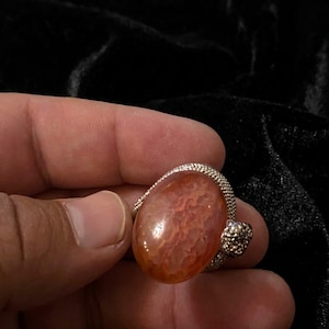 Puede incluir: Un anillo con una piedra naranja translúcida de forma ovalada y un patrón interno texturizado similar a una nube. La piedra está engastada en una banda de metal plateado con una textura fina, granulada o perlada en los bordes y el engaste.