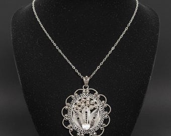 May include: Silver filigree pendant necklace. The pendant features a floral design with intricate details and a delicate chain. The necklace is displayed on a black mannequin, showcasing the craftsmanship and elegance of the jewelry.