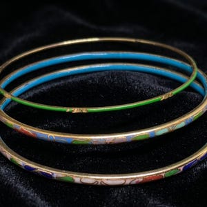 Puede incluir: Tres brazaletes finos de metal apilados. El brazalete superior es de color verde sólido con bordes dorados. Los brazaletes del medio y de abajo presentan un patrón floral de esmalte multicolor en tonos azul, verde, rojo y blanco con detalles en metal dorado.