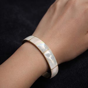 May include: A gold-toned bangle bracelet with rectangular mother-of-pearl inlays. The bracelet is worn on a wrist, showcasing the iridescent white and cream colors of the inlaid sections. The bracelet's design is simple and elegant.
