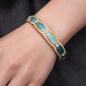 Può includere: Un braccialetto rigido dorato con accenti in smalto turchese e bianco. Il braccialetto presenta sezioni ovali in smalto con uno strass trasparente centrale. Il braccialetto è indossato al polso.