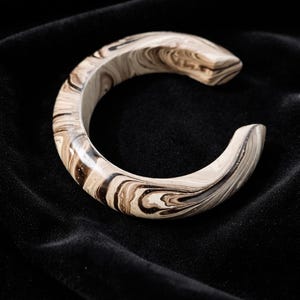 Può includere: Un bracciale a polsino effetto marmorizzato nei toni del marrone e del crema. Il bracciale aperto presenta un motivo a vortice, creando un'estetica unica e organica. Il bracciale è posto su uno sfondo nero, che ne evidenzia il design.