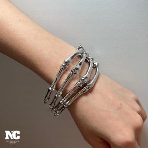 May include: A silver-colored bracelet shaped like a skeleton hand. The bracelet is worn on a wrist and features detailed bone structures. The jewelry has a gothic or alternative aesthetic. The brand logo "NC NOCTELLECHARM JEWELRY" is visible in the bottom left corner.