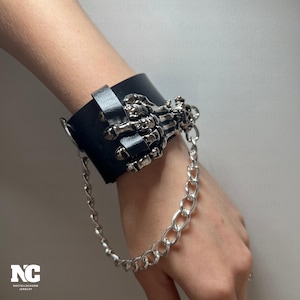 May include: Black leather wrist cuff featuring a silver skeleton hand design. The cuff is connected to a silver chain. The jewelry piece is a gothic accessory.
