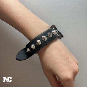 Puede incluir: Pulsera de cuero negro con tachuelas cónicas plateadas. La pulsera tiene un cierre de hebilla y se lleva en la muñeca. La joya es un accesorio de estilo punk o rock.