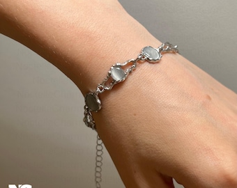 May include: A silver bracelet with oval, pale grey gemstones. The bracelet is linked by a fine chain and has an adjustable length. The gemstones are set in a decorative, openwork setting. The bracelet is shown on a person's wrist.
