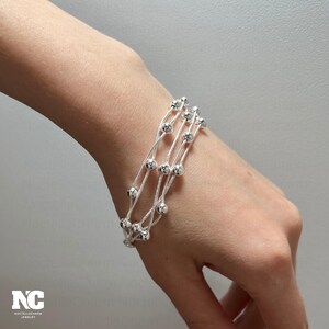 May include: A silver bracelet featuring three strands, each decorated with small, round beads. The bracelet is displayed on a wrist, highlighting its delicate design and reflective surface. This jewellery piece is a stylish accessory.