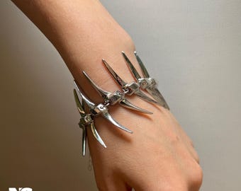 May include: A silver-toned bracelet featuring a series of stylized, pointed spikes. The bracelet is designed to wrap around the wrist, with each spike element connected by small links. The jewelry has a gothic or edgy aesthetic.