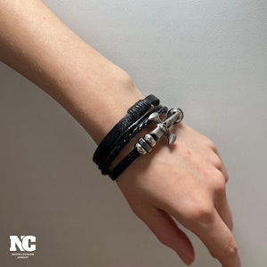May include: Three black leather bracelets are displayed. One is braided, another is wrapped, and the third has a silver anchor charm. The bracelets are stacked on a wrist, with a nautical design.