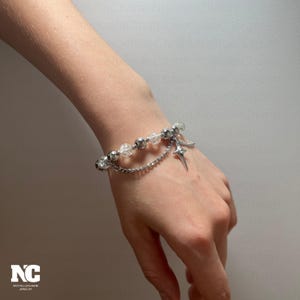 May include: A silver bracelet with clear, faceted beads and smaller silver beads. A silver chain dangles from the bracelet, holding a star-shaped charm. The bracelet is worn on a wrist.
