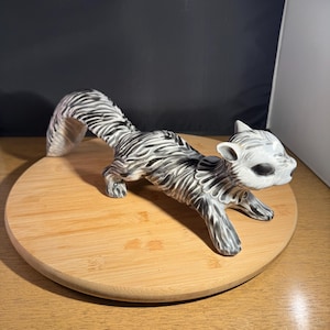 May include: A ceramic squirrel figurine in a crawling pose. The squirrel is painted in shades of gray and white with a striped pattern. It is displayed on a round wooden surface.
