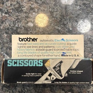 May include: A Brother automatic electric scissors product box featuring text that highlights features such as a guide light for patterns, a blade guard, high carbon steel blades, and a contoured shape for either hand. The box is made in the U.S.A.