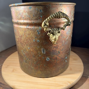 May include: A vintage copper bucket with a tarnished finish, featuring a decorative brass handle with a face design. The bucket sits on a round wooden base, showcasing its aged patina and unique aesthetic.