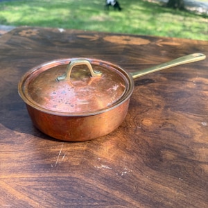 May include: A small copper saucepan with a lid and a long brass handle. The pan is round with a slightly curved body. The lid has a brass handle. The pan is sitting on a wooden surface.