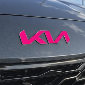 May include: A dark gray vehicle hood featuring a bright pink Kia logo emblem. The emblem is centered on the smooth, metallic surface above the black front grille, displaying the stylized lettering of the brand in a vibrant, solid pink color.
