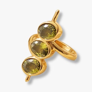 May include: A gold ring with three oval, olive-green gemstones. The ring has a distinctive design, with the gemstones arranged vertically and a delicate band. The ring is set against a white background.