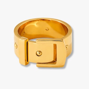 Könnte beinhalten: Ein goldfarbener Ring mit Schnallendesign. Der Ring hat eine rechteckige Schnalle mit abgerundetem Stift und kleinen Nieten. Der Ring besteht aus glänzendem, reflektierendem Metall.