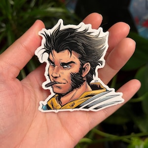 Puede incluir: Una pegatina con una ilustración de estilo cómic de un hombre con pelo negro, barba y un cigarro. El hombre lleva un atuendo amarillo y blanco. La pegatina se sostiene en una mano.