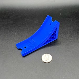 Op de afbeelding: Een helderblauwe, gebogen plastic object met een honingraatpatroon aan één kant. Het heeft twee kleine gaten en een inkeping. Een munt ligt ernaast ter schaal. De lengte is ongeveer 7,6 cm.