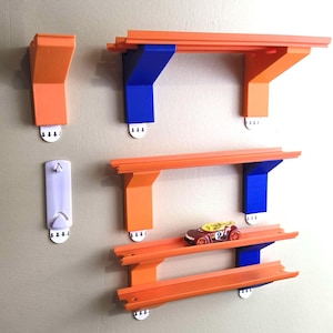 Puede incluir: Estanterías naranjas y azules para pistas de coches de juguete montadas en una pared. Las estanterías están diseñadas para guardar coches de juguete, con una estantería que contiene un coche de juguete rojo y amarillo. Incluye ganchos adhesivos blancos.