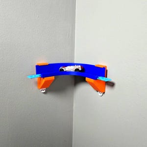 Könnte beinhalten: Eine blaue und orangefarbene, eckmontierte Spielzeugauto-Rennstrecke mit einem kleinen Spielzeugauto. Die Rennstrecke ist für eine Ecke konzipiert und hat ein gebogenes Design. Das Spielzeugauto ist weiß mit roten und schwarzen Details.