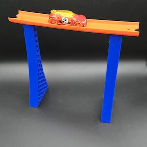 Puede incluir: Un coche de juguete rojo con el número 2 está sobre una pista naranja sostenida por pilares azules. La pista está elevada, creando una estructura similar a una rampa. El fondo es gris oscuro.