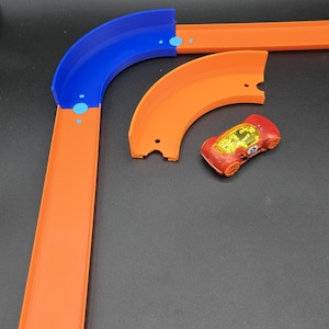 Op de afbeelding: Een oranje en blauw speelgoed racebaan met een rode raceauto. De baan heeft gebogen en rechte secties. De raceauto is rood met oranje wielen en een gele binnenkant. De baan is gemaakt van plastic.