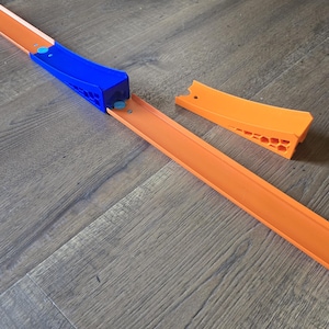 Op de afbeelding: Een speelgoed autoracebaan set in oranje en blauw. De baan is oranje, met een blauw lanceermechanisme en een extra oranje stuk. De set is ontworpen voor speelgoedauto's en ligt op een houten vloer.