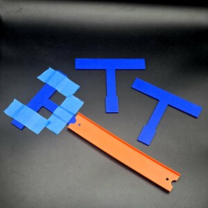 Op de afbeelding: Een oranje speelgoedautoracebaan met blauwe tape en drie blauwe T-vormige stukken. De baan is schuin en de stukken liggen verspreid op een zwart oppervlak. Het beeld suggereert een constructie- of montagethema.