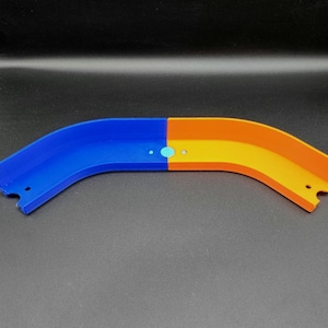 Op de afbeelding: Een gebogen plastic speelgoedbaanstuk, verdeeld in blauwe en oranje secties. De baan heeft kleine cirkelvormige details en is ontworpen voor speelgoedauto's. Het is geplaatst tegen een donkere achtergrond.