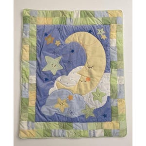 May include: A baby quilt featuring a whimsical night sky design. The quilt has a blue center with a yellow crescent moon and smiling stars. The border is a patchwork of green, yellow, and blue squares.
